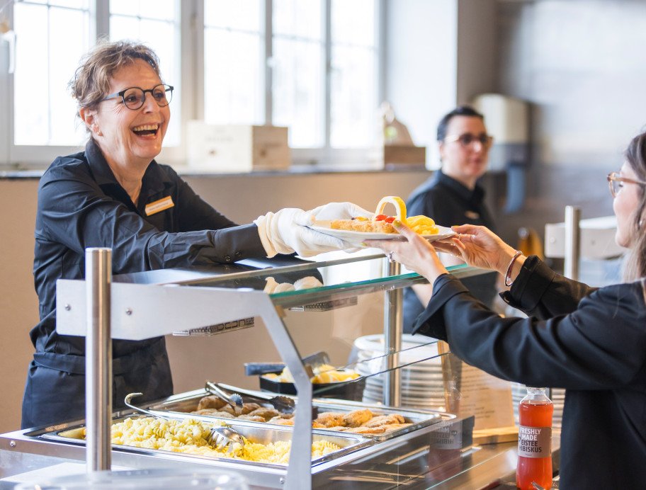 Mitarbeitende übergibt einen Teller mit Essen an einen Gast